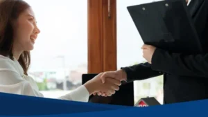 Agente y cliente se dan la mano tras finalizar la negociación de casas en venta. Modelo de casa y documentos en la mesa.